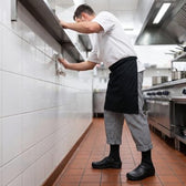 Male cook in a white chef jacket, checkered pants, black Shift Socks and black kitchen shoes wiping down a tiled wall in a restaurant kitchen.
