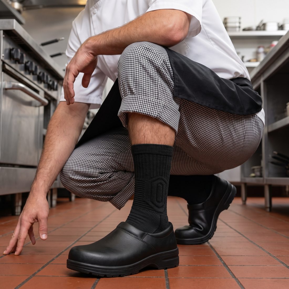 Cook crouching in a restaurant kitchen, black Shift Sock sitting snug above a black kitchen shoe.