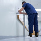 Male nurse in navy scrubs, black Shift Socks and white clogs restocking a glove dispenser in a hospital corridor.