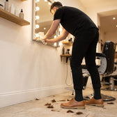 Barber in black, wearing black Shift Socks and tan leather shoes, wiping a light-bulb mirror in a hair salon.
