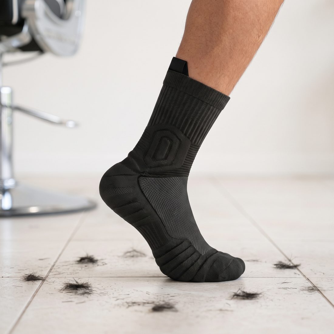 Man's foot on tiptoe in a black Shift Sock on a salon floor, with hair clippings and a barber chair behind.