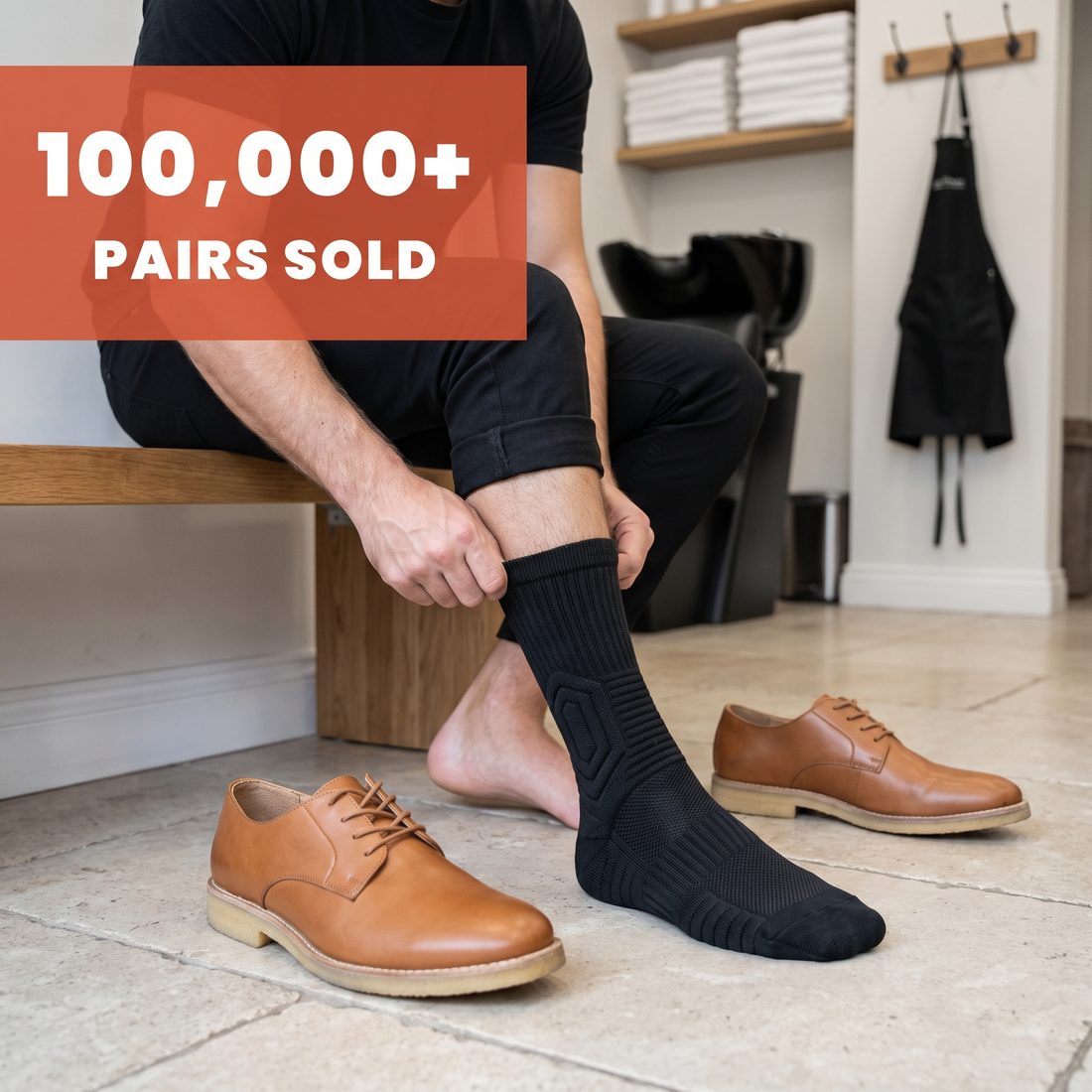 Barber on a back-room bench pulling on a black Shift Sock, tan leather shoes beside him, 100,000+ pairs sold.