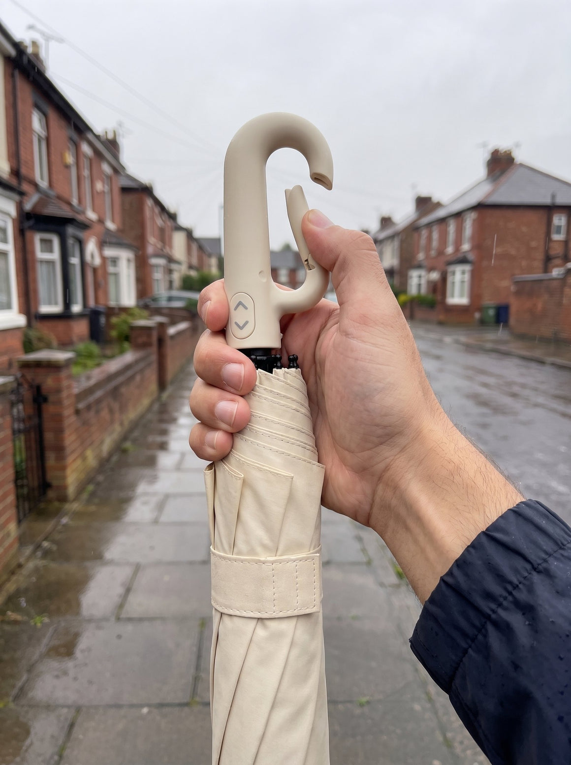 A man's hand opening the carabiner clip on the handle of a folded beige umbrella, on a wet terraced street