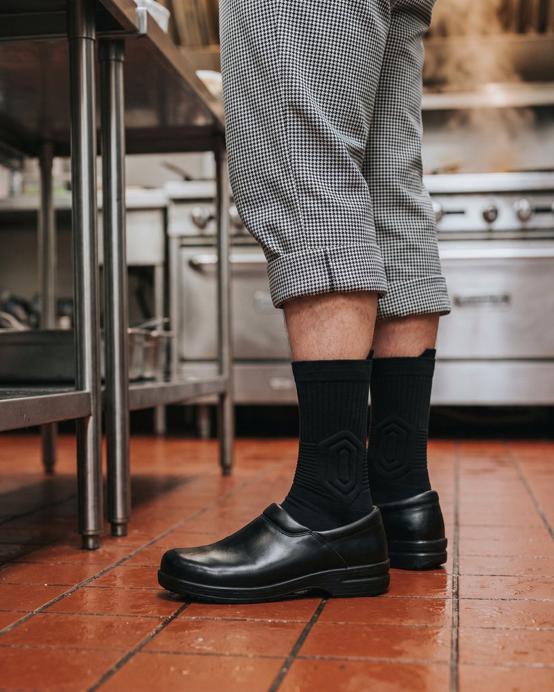 Legs of a cook in checkered pants, black Shift Socks and black kitchen clogs on a wet tiled kitchen floor.