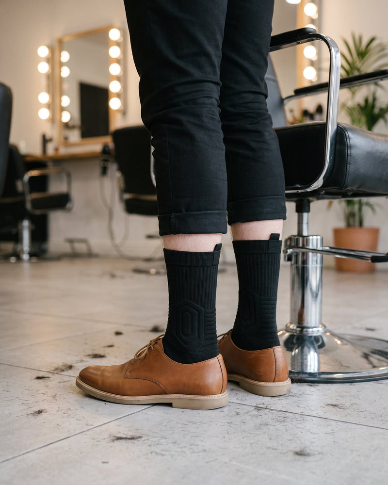 Legs of a hairstylist in black pants, black Shift Socks and tan leather shoes next to a salon chair.