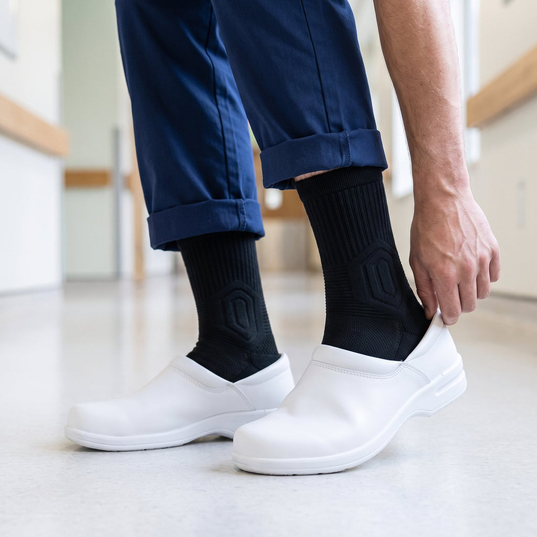 Male nurse in navy scrubs adjusting a white hospital clog over black Shift Socks on a hospital floor.