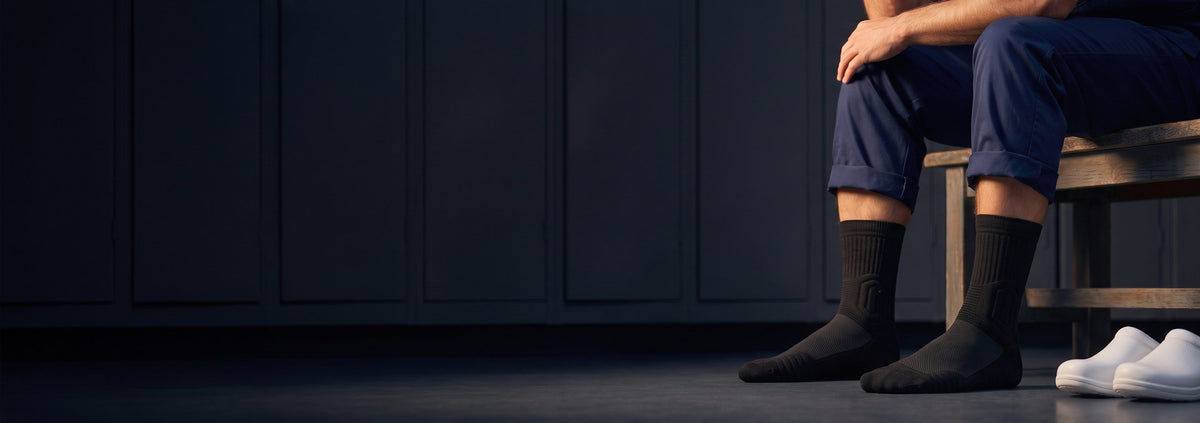 Male nurse sitting on a locker room bench after a shift, in black Shift Socks, white clogs beside him.